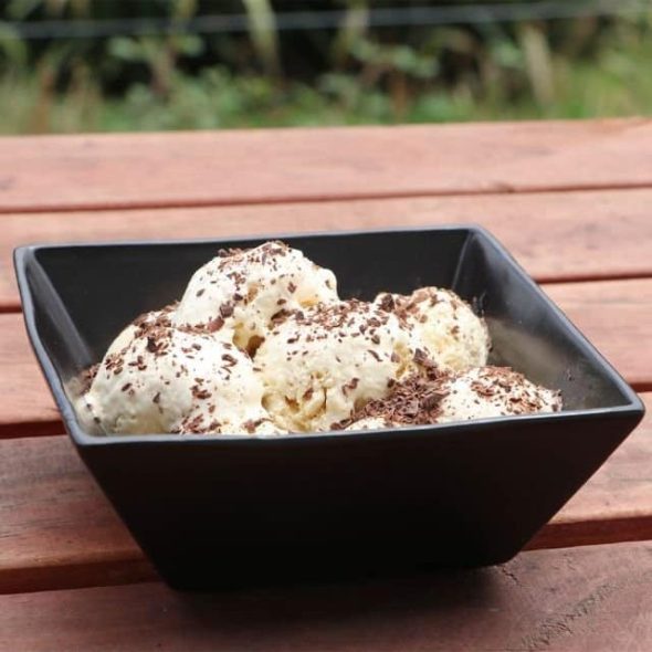 A dark square bowl filled with scoops of creamy Tawari Honey Ice Cream, generously sprinkled with chocolate shavings, resting on a rustic wooden surface outdoors.