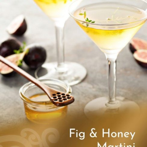 A close-up of two elegant martini glasses filled with a golden Fig & Tawari Honey Martini, garnished with a sprig of thyme. Fresh figs and a small jar of honey with a honey dipper are artfully arranged nearby.