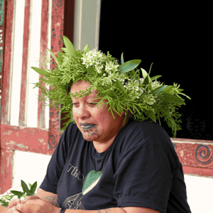 Māori Cultural Heritage: Woman with Tāwari Flower Lei Po'o Māori woman, Kathy Taputu, wearing a beautiful head wreath (lei po'o) made of native Tāwari flowers and foliage. She has a traditional chin tattoo (moko kauae) and is seated in front of a carved marae, representing the deep cultural connection to the whenua (land) in New Zealand.