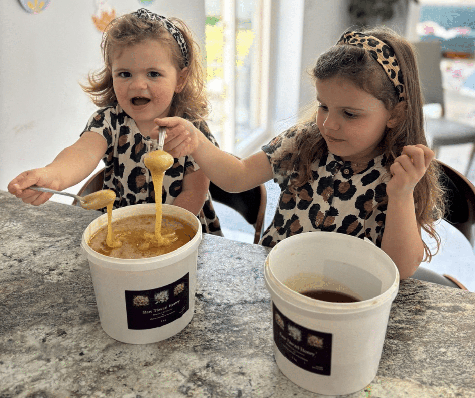 Kids Enjoying Raw Tawari Honey from Manawa Honey NZ - A Sweet & Healthy TreatKids Enjoying Raw Tawari Honey from Manawa Honey NZ - A Sweet & Healthy Treat Two adorable little girls in matching leopard print outfits happily eating raw Tawari honey with spoons directly from large tubs, showing their love for Manawa Honey NZ's natural product.
