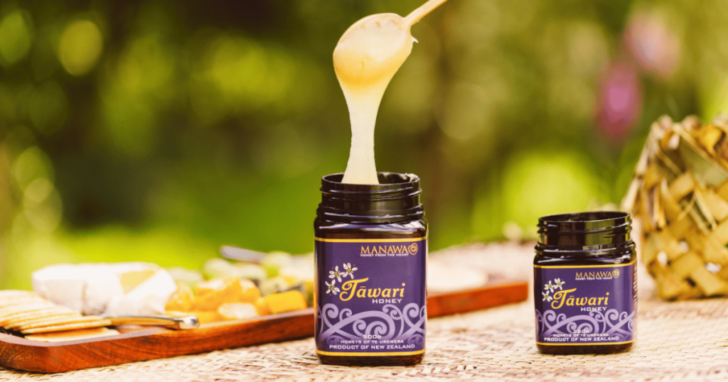 Manawa Honey NZ Tawari Honey - Premium New Zealand Honey | Sweet & Creamy Close-up of creamy Manawa Honey NZ's Tawari Honey being spooned from a dark purple jar, with a smaller 250g jar of Tawari honey and a cheese board in the blurred background. Premium New Zealand honey known for its sweet, buttery taste and light color.