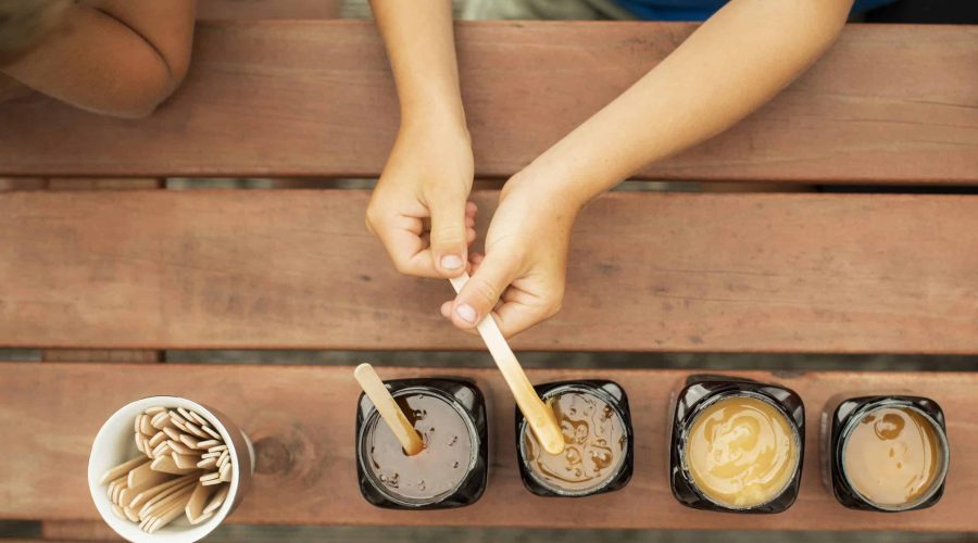 Top-down view of a person using wooden sticks to taste four different varieties of Manawa honey in dark jars on a wooden table, including Mānuka, Tāwari, Pua-a-Tane and Rewarewa.