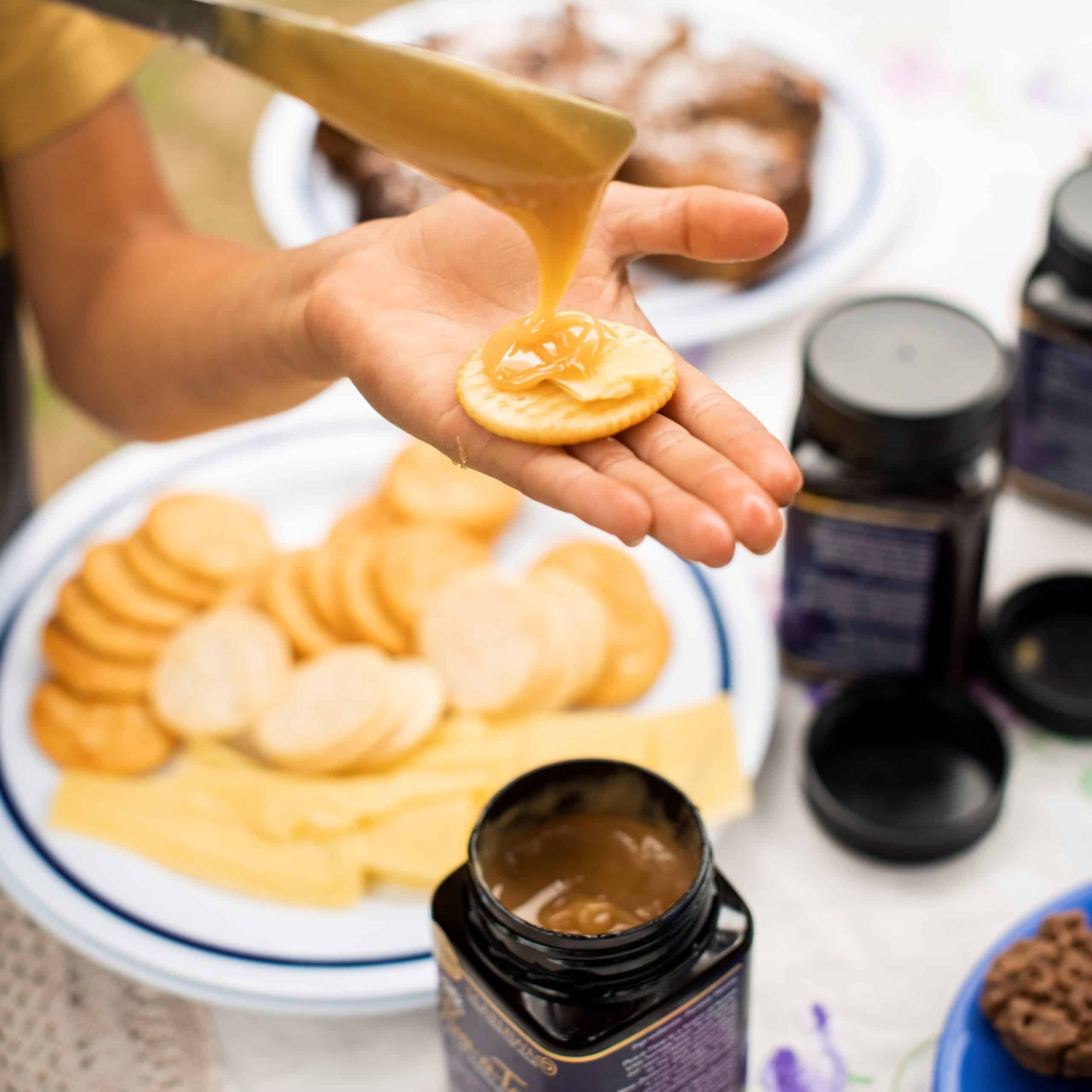 A close-up of thick golden Manawa honey being drizzled from a knife onto a cracker with cheese, held in a hand over a platter of snacks.